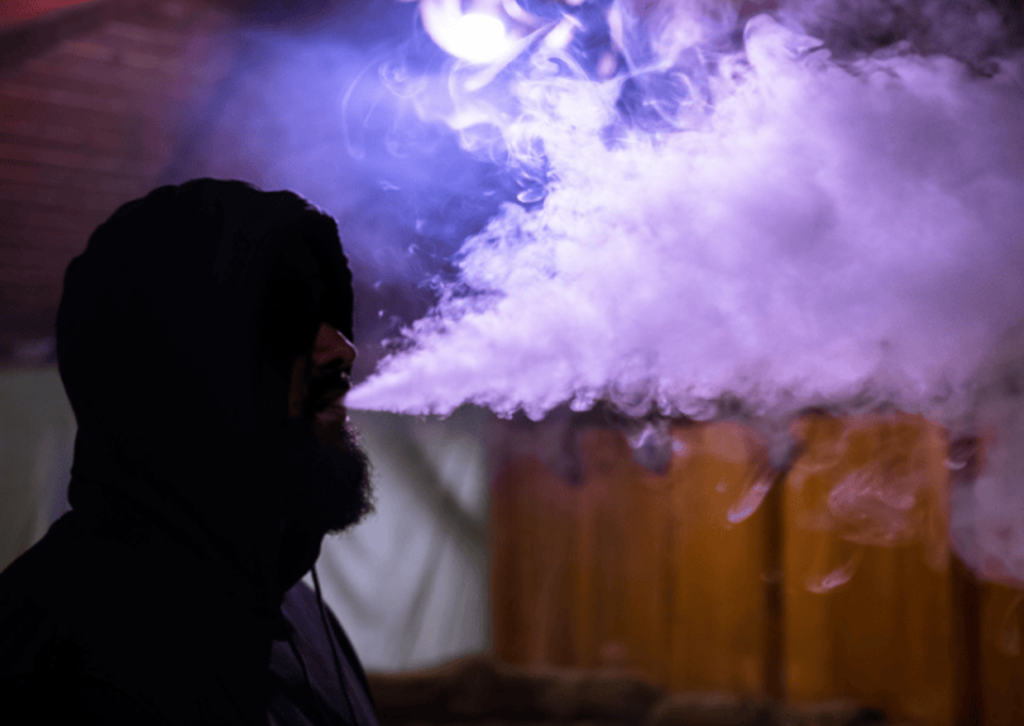 Man sitting in dark room with purple light blowing vape smoke