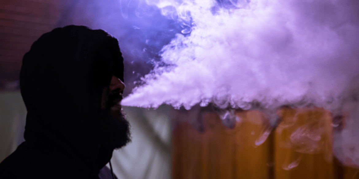 Man sitting in dark room with purple light blowing vape smoke