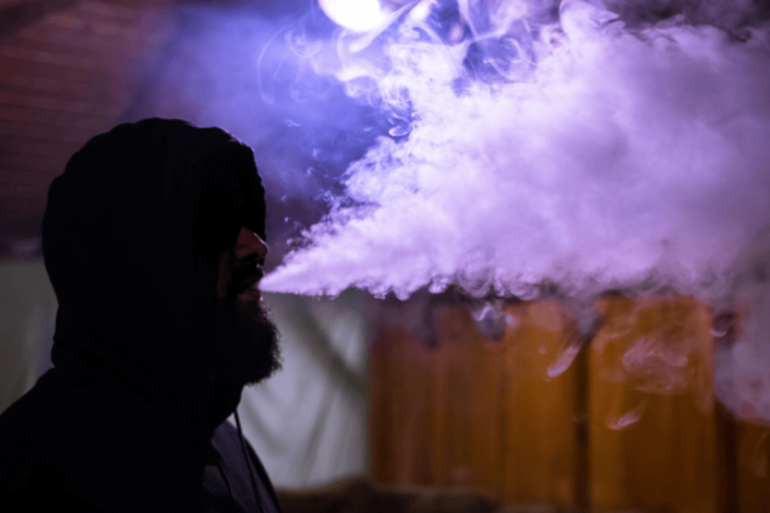 Man sitting in dark room with purple light blowing vape smoke